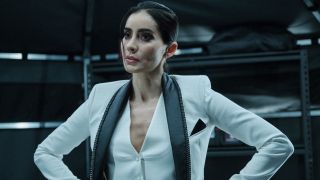 Paola Núñez stands with her arms akimbo, in a white suit with black scarf in Resident Evil.