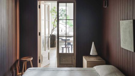 A small bedroom painted in a dark maroon and dark gray color. There is minimal art on the walls and the bedding is a thin, minimal, linen style. There are two wooden doors with windows leading into a bright living room