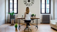 Woman sitting at a desk with a clock on the wall in front of her. She is seen from behind. She is in a domestic setting. 