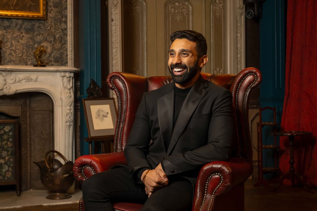 Jaz Singh sits in a red leather armchair in the Traitors castle, wearing a black suit. He is smiling, and looking off to the left of the frame.