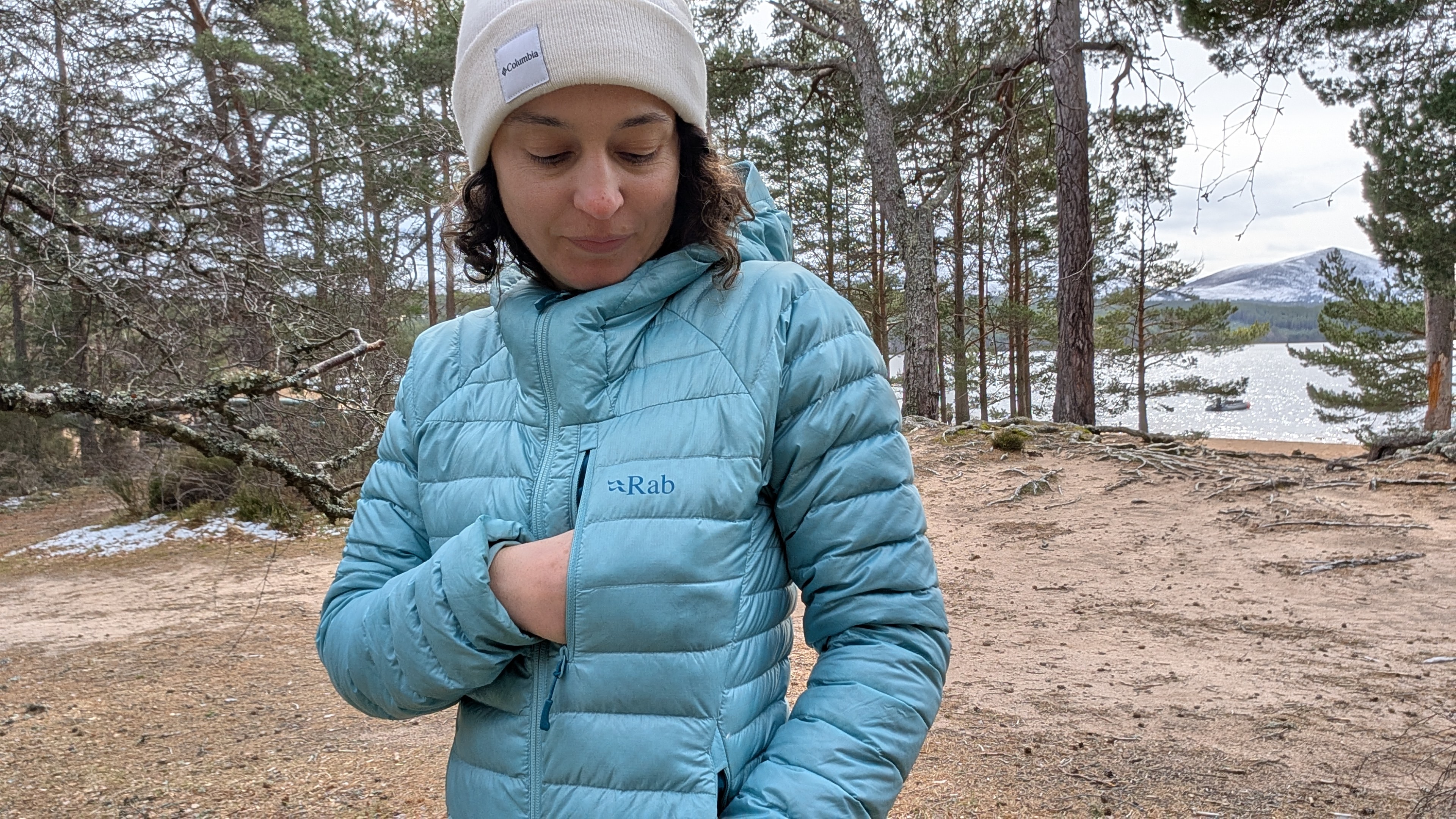A woman on a beach near a lake opening the chest pocket on a bright blue down jacket