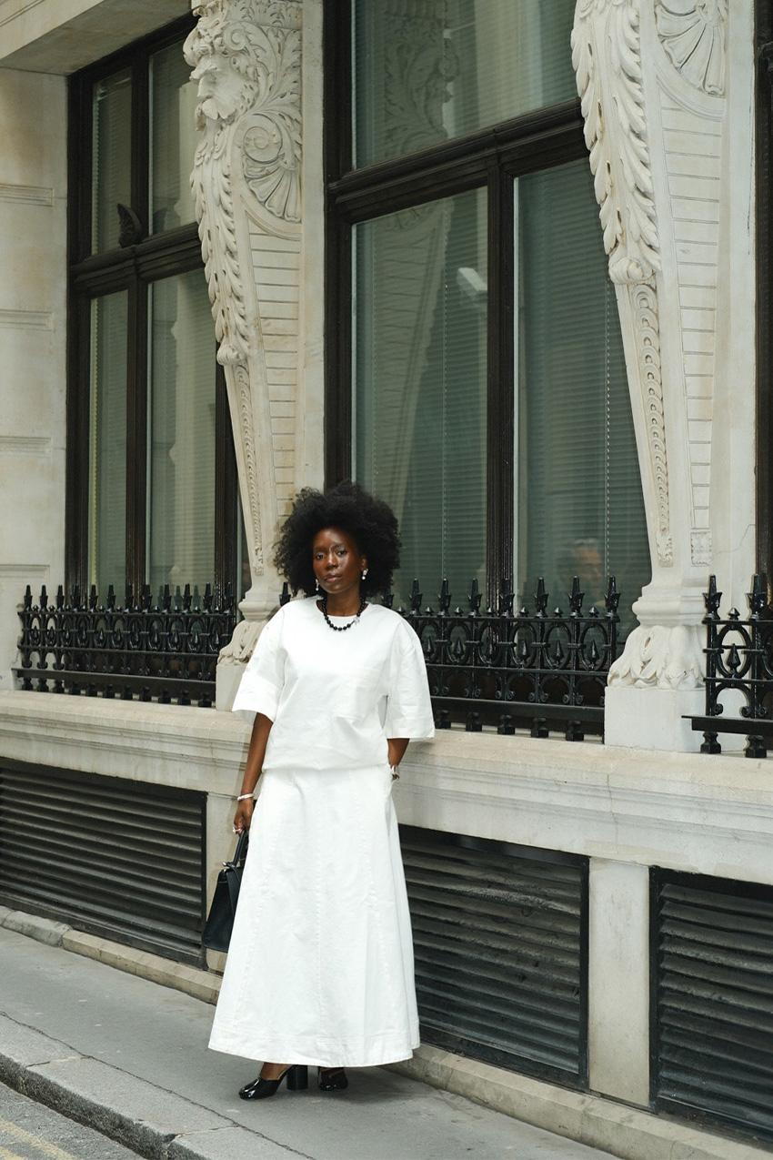White outfit idea for summer is shown in a photo of a woman standing outside on the sidewalk wearing a black necklace with an oversized white t-shirt, a white maxi skirt, black heels, and a black handbag