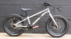 The Early Rider Seeker 16, with its polished silver finish, all black componentry, and Red Funn pedals, against the backdrop of a black painted breeze block wall 