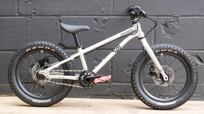 The Early Rider Seeker 16, with its polished silver finish, all black componentry, and Red Funn pedals, against the backdrop of a black painted breeze block wall 