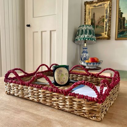 Woven seagrass tray with red wavy trim holding a candle in front of lamp and bowl of fruit