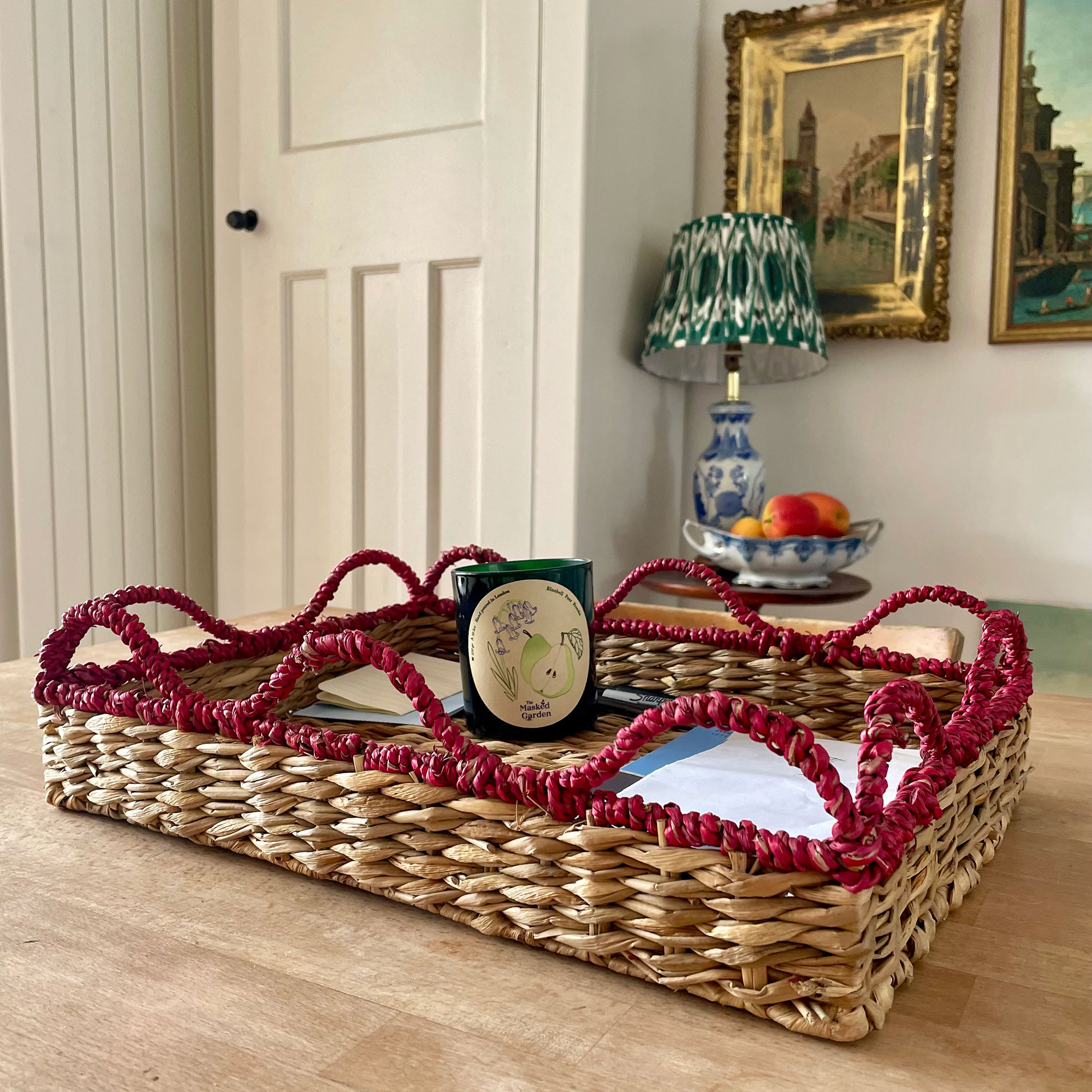 Woven seagrass tray with red wavy trim holding a candle in front of lamp and bowl of fruit