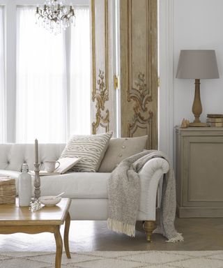 A living room with white sofa, taupe cushions and throw, and gold Rococo-style door