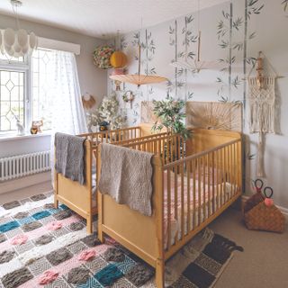 Two matching wooden cribs in front of green patterned wall, with cosy rug and blankets