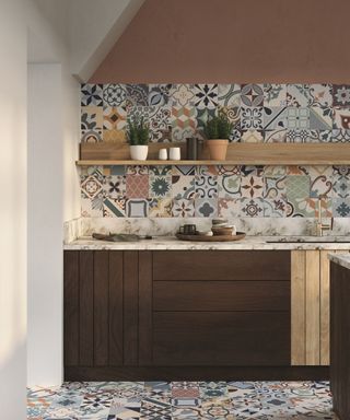 A kitchen with a white wall and open doorway to the left, a dark brown cabinet with white countertops, a wooden wall shelf with plants on it, and colorful blue tiles on the walls and floor