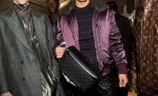 Close up view of male models wearing looks from Louis Vuitton's collection. One model is wearing a grey metallic piece with grey coat over the top. Next to him is a model wearing a dark purple striped jumper, purple jacket with fur and his black trousers and bag feature the signature Louis Vuitton pattern. Another model is wearing a brown jacket with the signature Louis Vuitton pattern. And in the background there are two models wearing black pieces