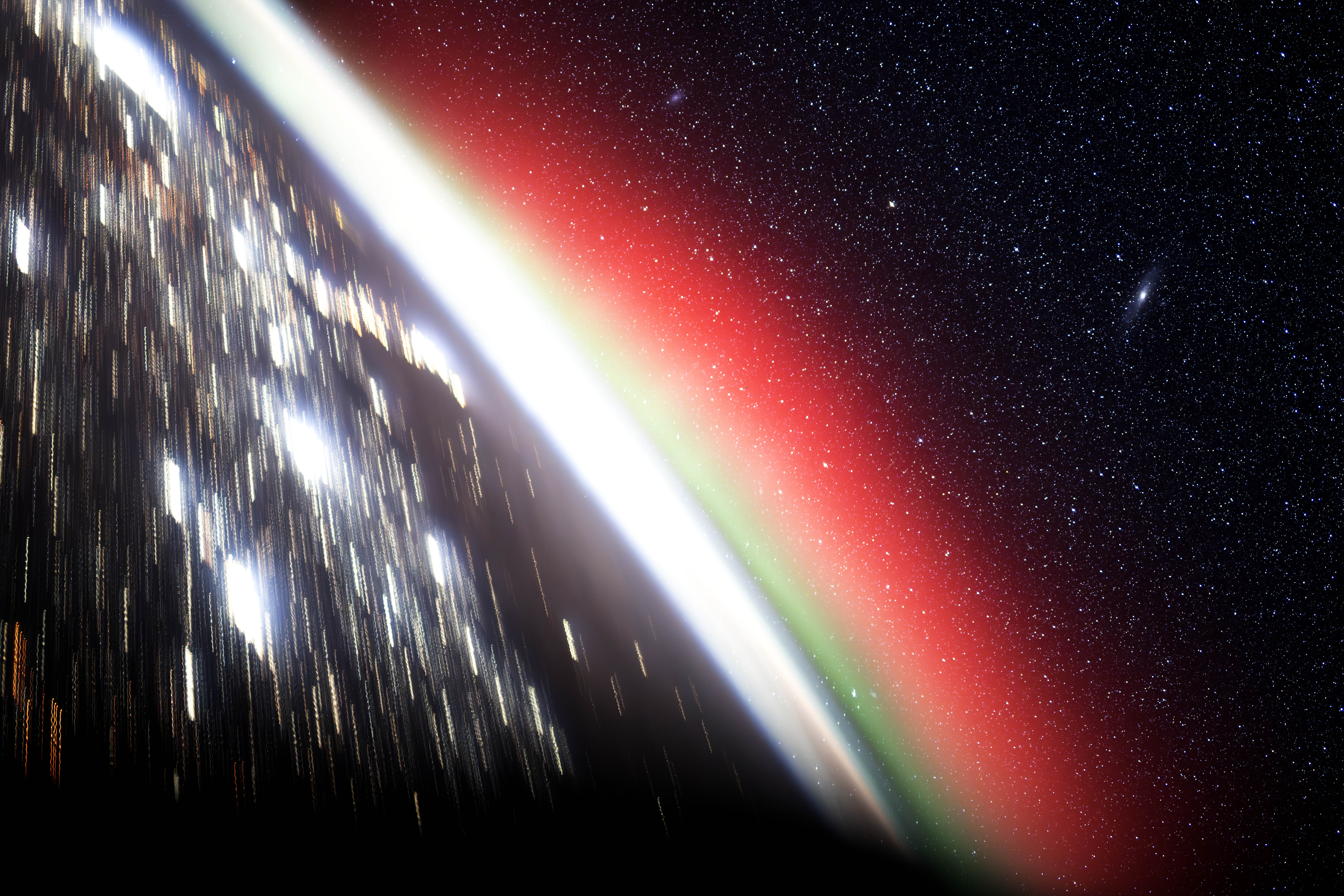 Earth pictured from space by astronaut Don Pettit. The lights on the planet&#039;s surface are blurred by its axial motion, while the stars beyond are pin-sharp against the blackness of space. The atmosphere is shown as a blown out white curve, with a nebulous red haze beyond it.