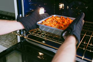 Placing a tray of food into the oven with oven gloves on.