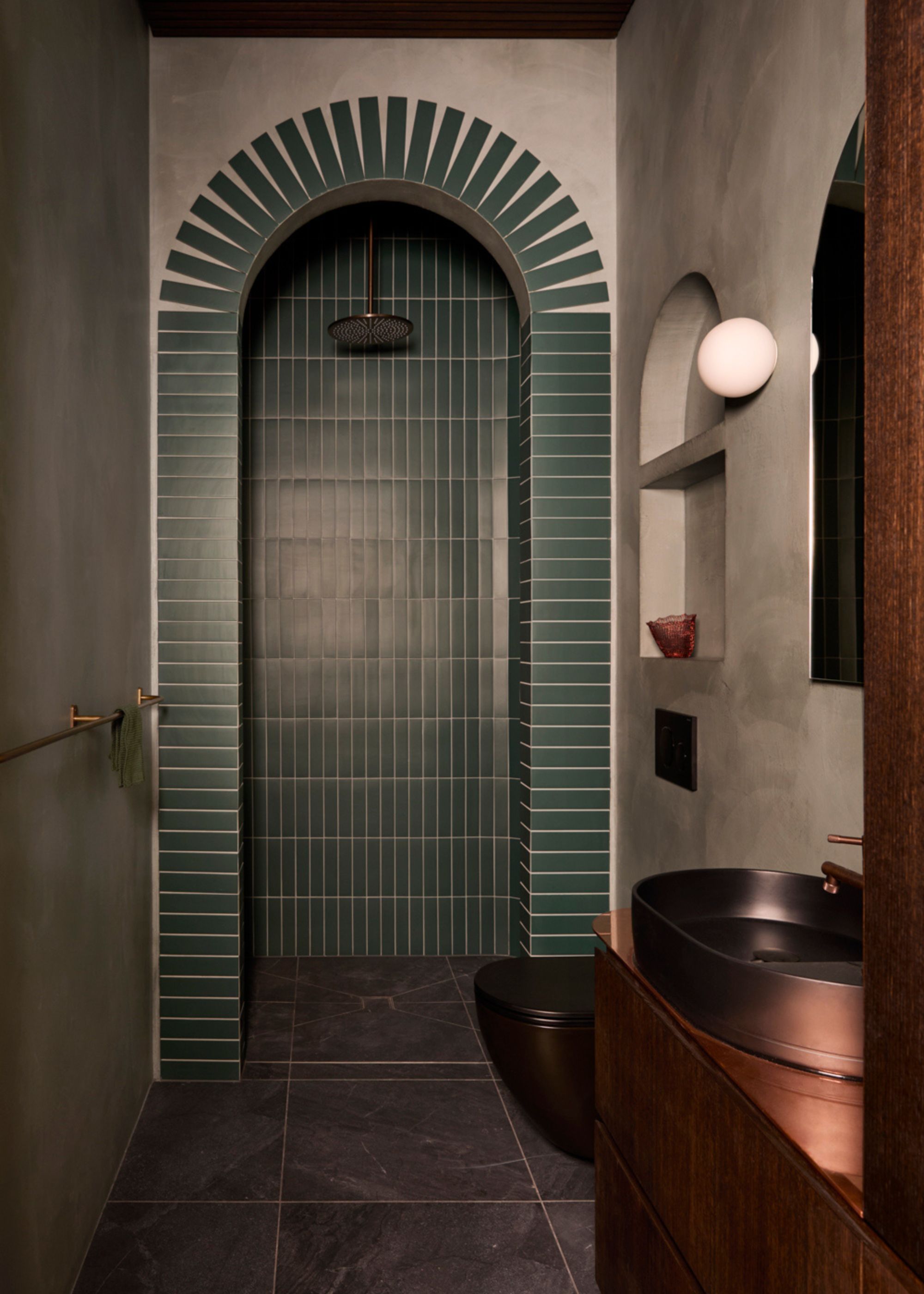 A bathroom with an arched shower space, an arched wall mirror, a round sconce, and a brass-y basin