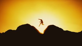 Silhouette of a person jumping between two rocks against a vibrant orange sunset sky, capturing a moment of freedom and adventure