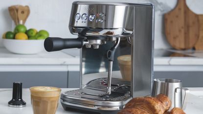 Image of a silver Sage Bambino Plus coffee machine in a white and gray kitchen with a latte and croissants around it.