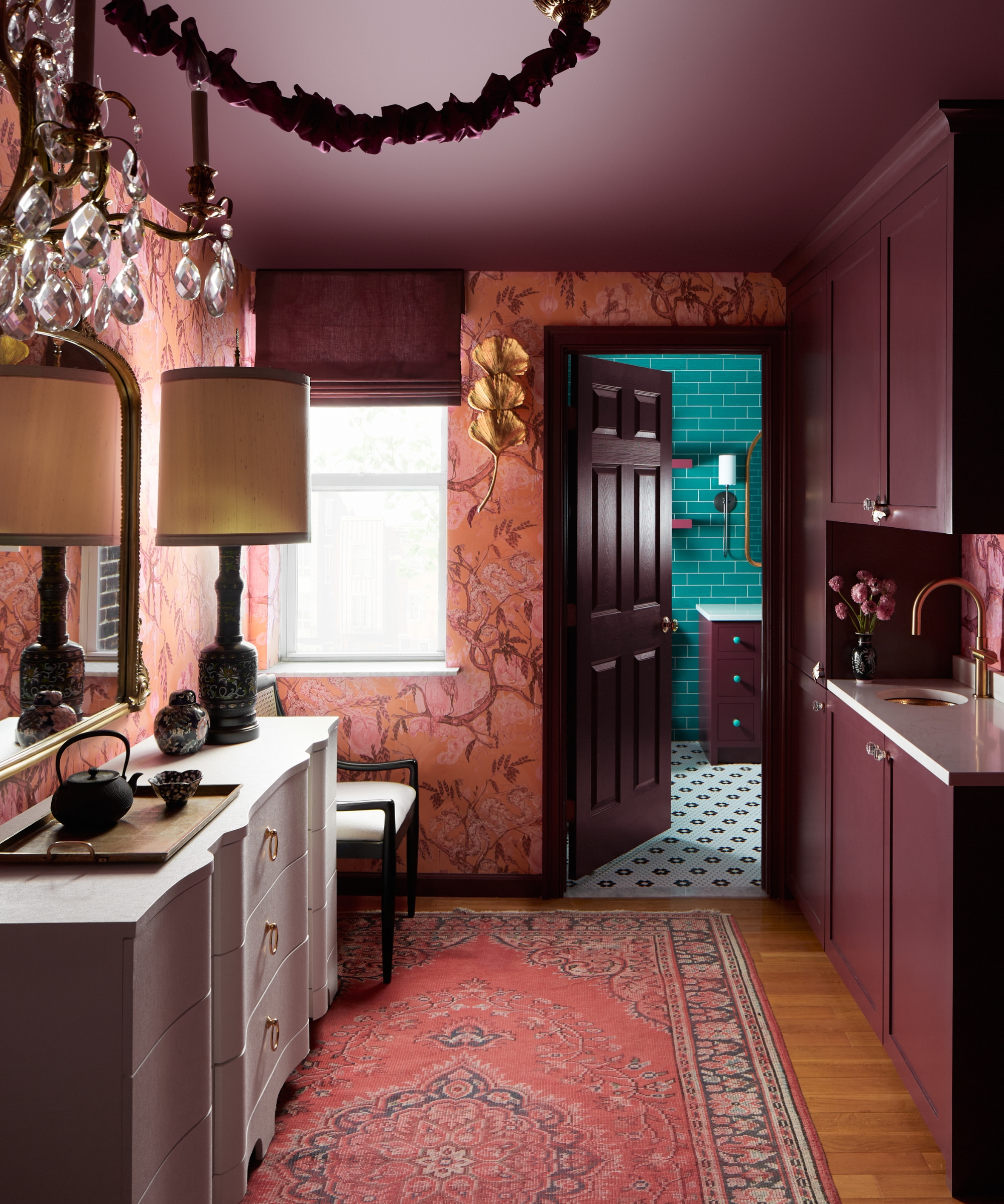 Maroon cabinetry and ceiling with pink wallpaper, antique rug, brass accents, and a teal-tiled bathroom beyond in a richly layered interior.