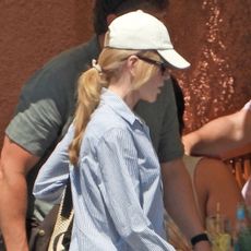Nicole Kidman in Portofino wearing a white baseball cap, blue-and-white-striped button-down shirt, white cut-off denim shorts, a Loewe raffia shoulder bag, and white leather Adidas Samba sneakers
