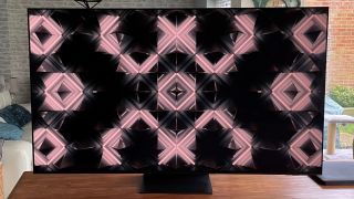 The Samsung S95C OLED TV showing abstract pink and black geometric pattern on wood table.