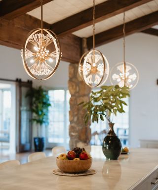 A marble kitchen island with a wooden fruit bowl and plants on it, three circular brass pendant lights above it, and dark wooden beams on the ceiling