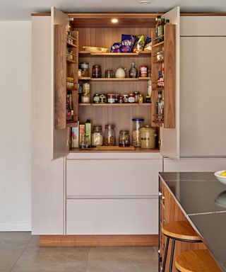 A pale pink kitchen cabinet with open doors with wooden shelving on the doors and inside containing jars, crisps, jams, and spices, with a black kitchen island with wooden stools in front