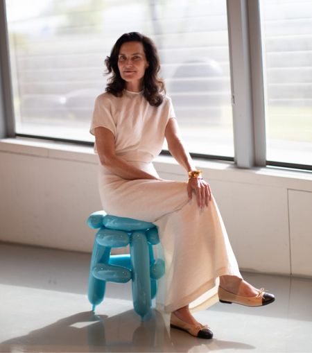 A woman dressed in a pale pink maxi dress with short sleeves sits on top of an inflated-looking stool in pale blue in a sun-filled gallery.