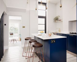 Open plan kitchen with blue fitted units and kitchen island on white painted floorboards, wood and copper bar stools.