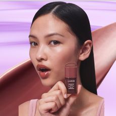 A model with black hair holding up a laneige lip tint. She is in front of an oversized swatch of the product, which is overlaid over a lavender, textured background.