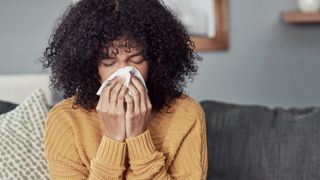 A woman is shown sat on a couch. She is blowing her nose with a tissue. 