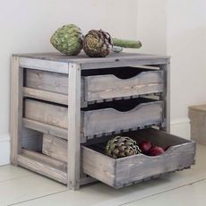 A small wooden unit in distressed pale blue with three drawers, one of which is open revealing vegetables within.