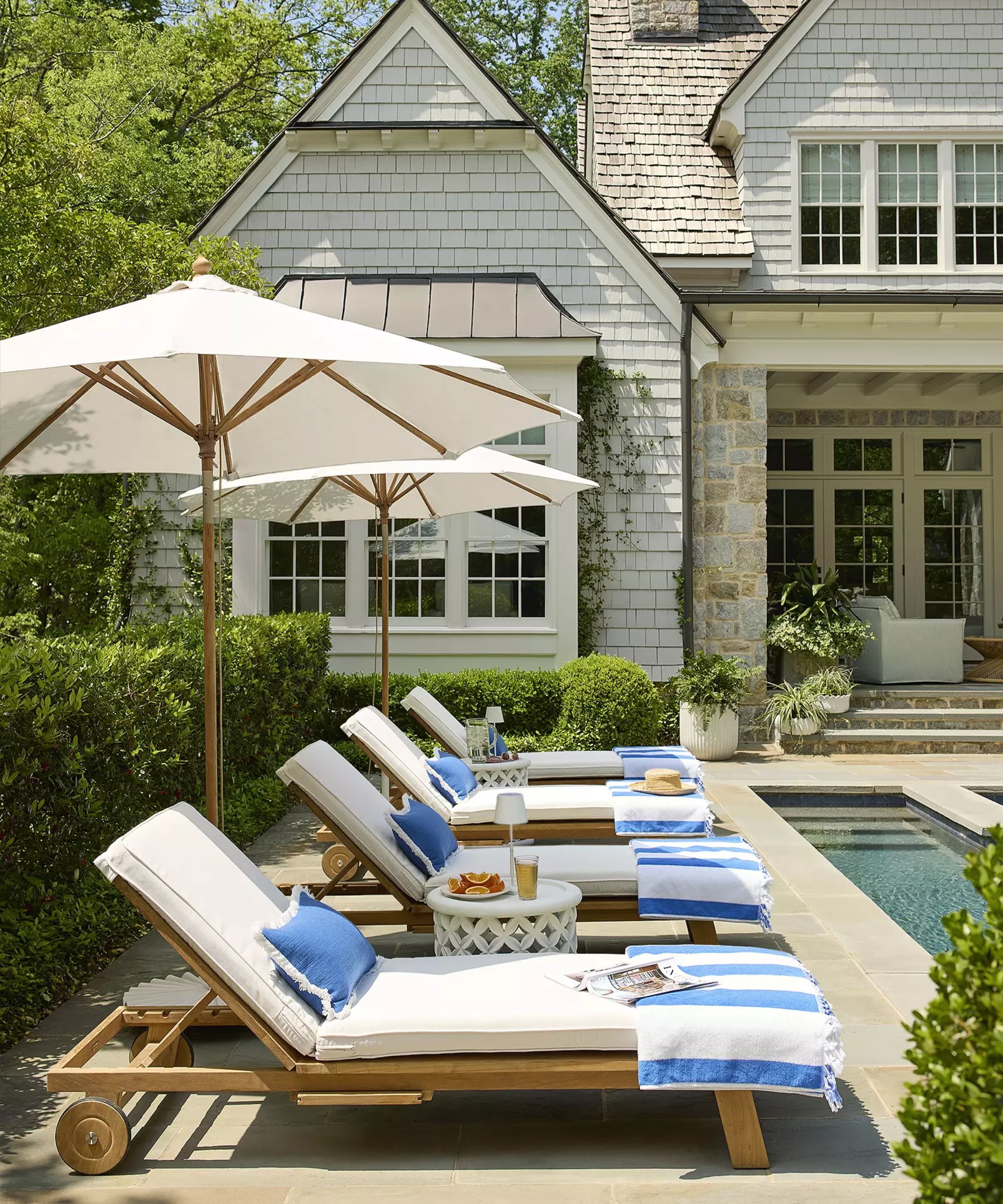 hamptons white and stone house with hegdges and a large pool with teak sun loungers with white cushions and white patio umbrellas with blue pillows and striped towels