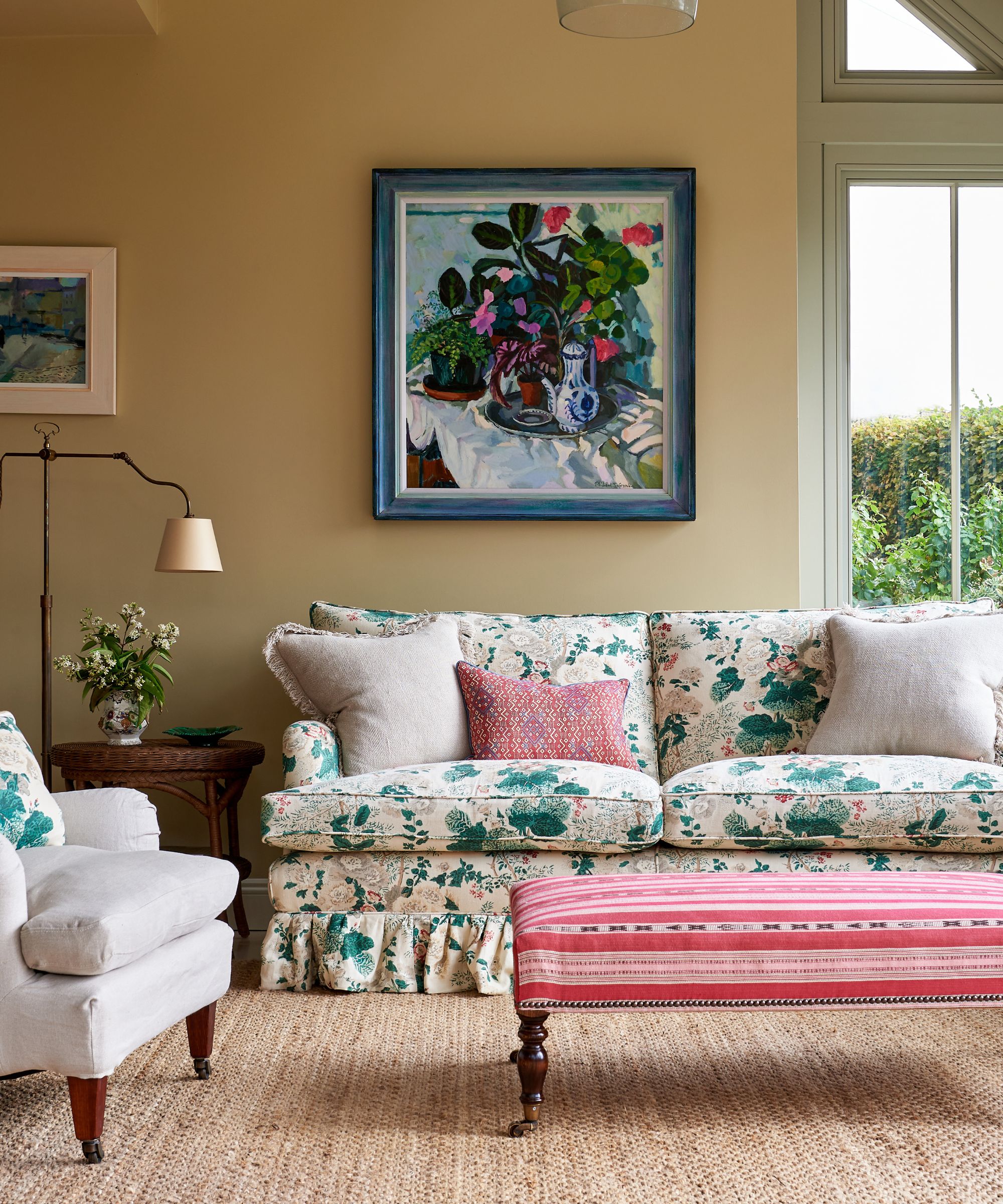 Country living room with pale yellow walls, abstract still life of a vase with flowers on the wall, floral skirted sofa, pink striped ottoman, cream armchair and bronze floor lamp with side table beneath it