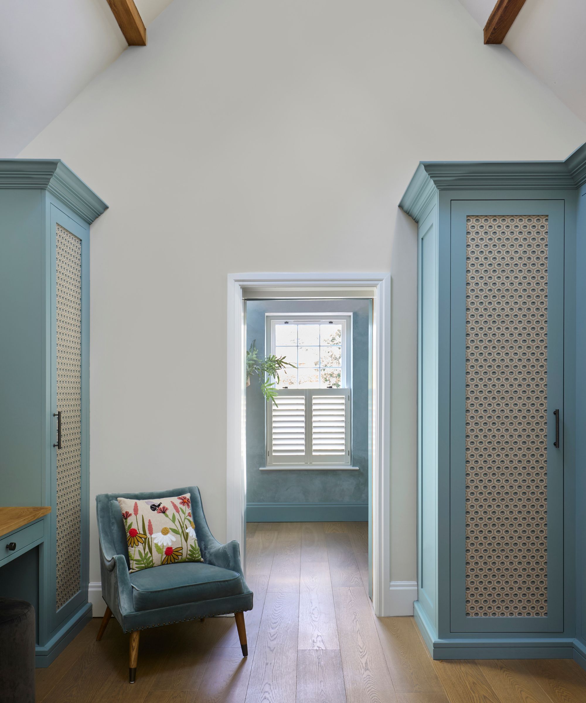 A loft dressing room with high white walls and exposed wooden beams. Two tall teal wardrobes with patterned inset panels stand opposite each other. Between them, a soft blue armchair with a floral cushion sits on warm wood flooring.