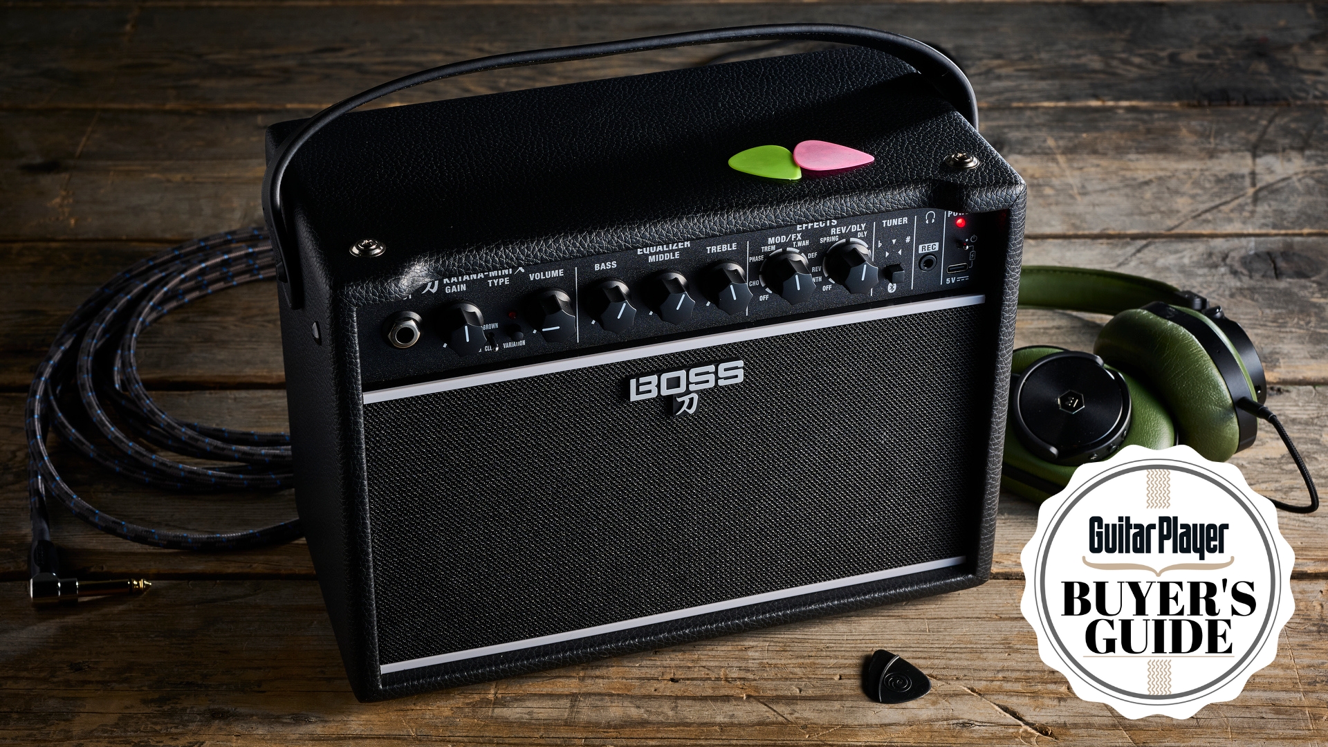 A Boss Katana Mini X mini guitar amp on a wooden floor with two guitar picks on top of it. In the background lies a guitar cable and a pair of headphones.