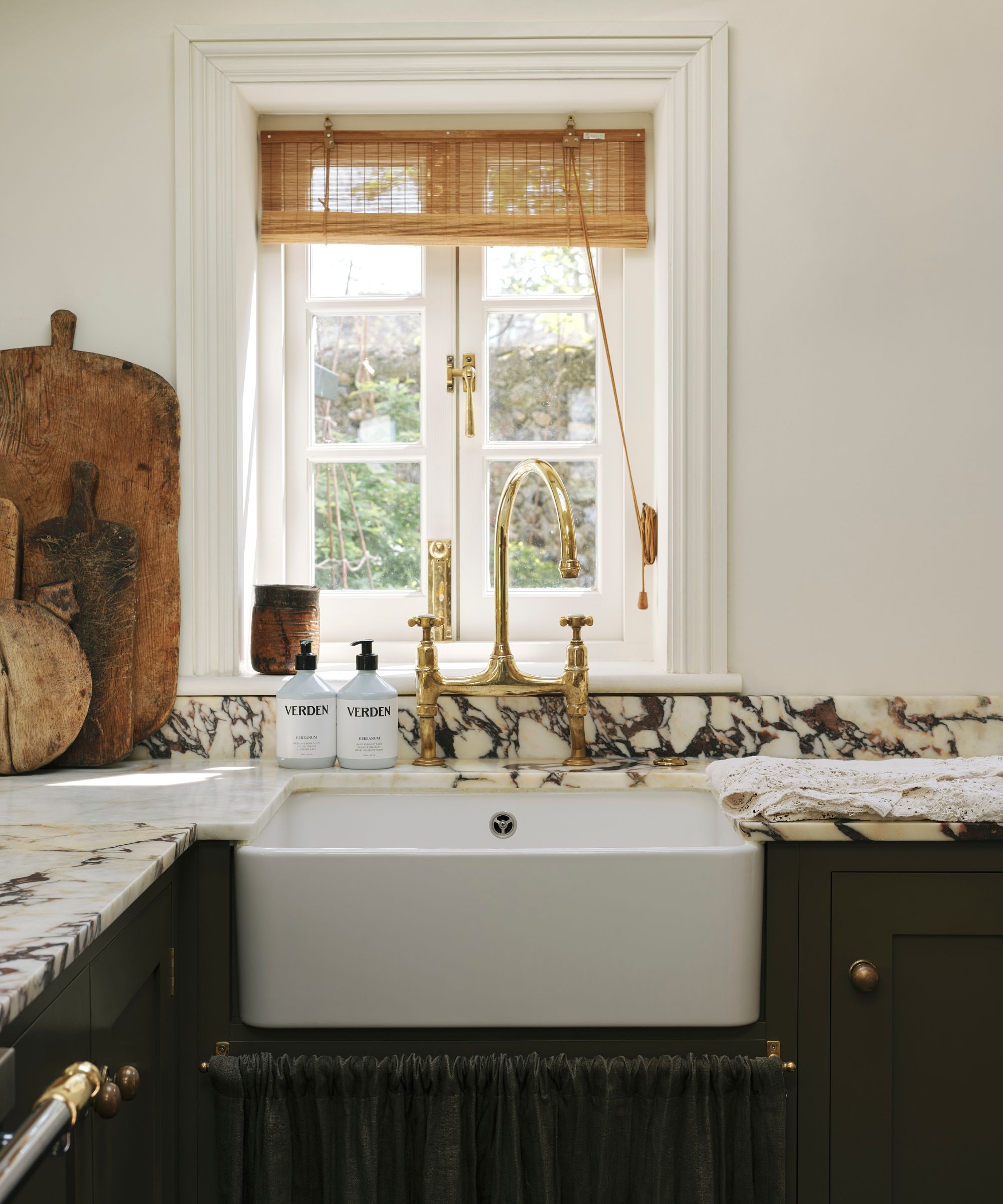 An olive green kitchen with warm white walls, marble countertops, a matching olive green sink skirt, and a seagrass blind over the window