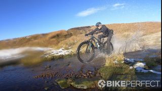 Guy Kesteven rides his bike through a river crossing, and water is splashing towards the camera