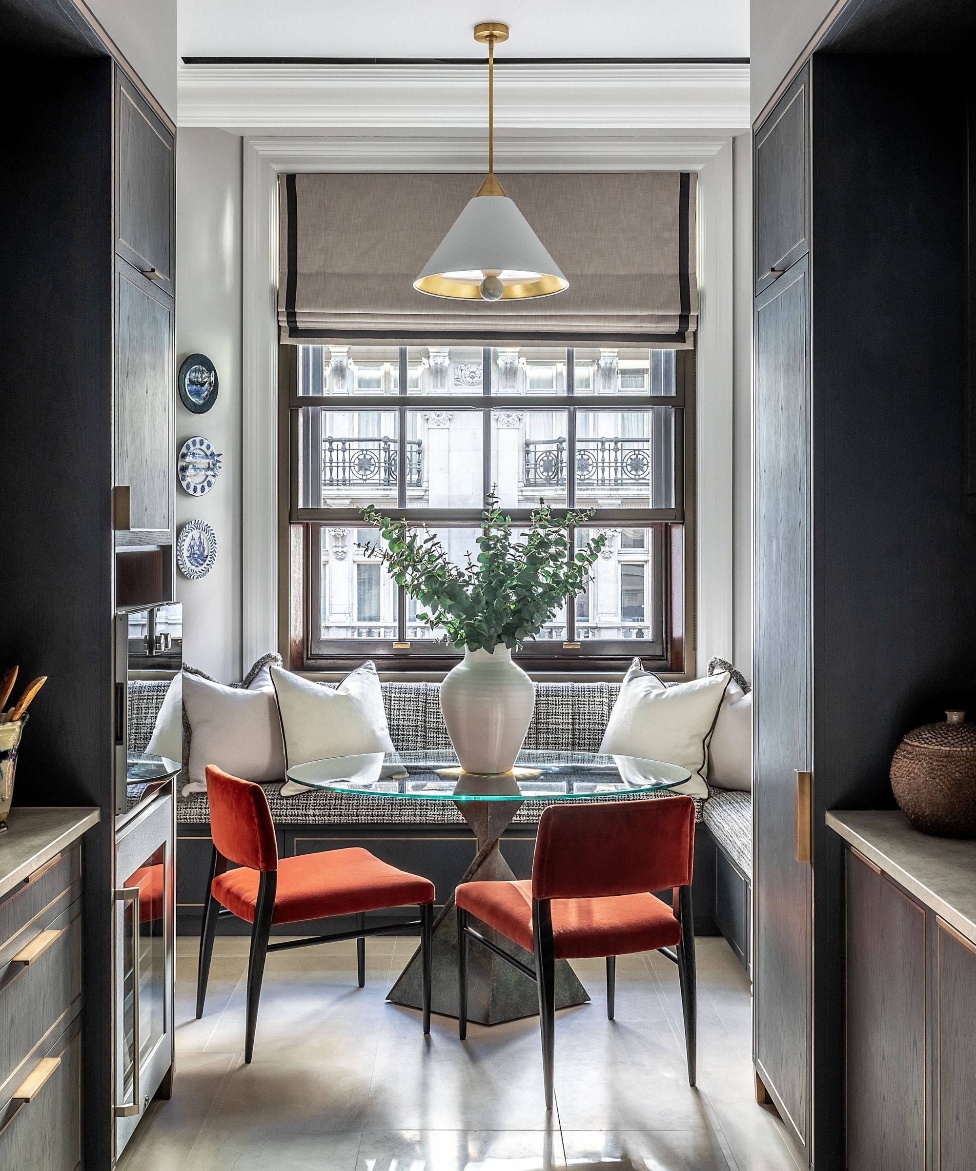 A small round glass kitchen dining table in front of a banquette bench, with two red velvet chairs tucked beneath. In a large window at the end of a narrow kitchen.