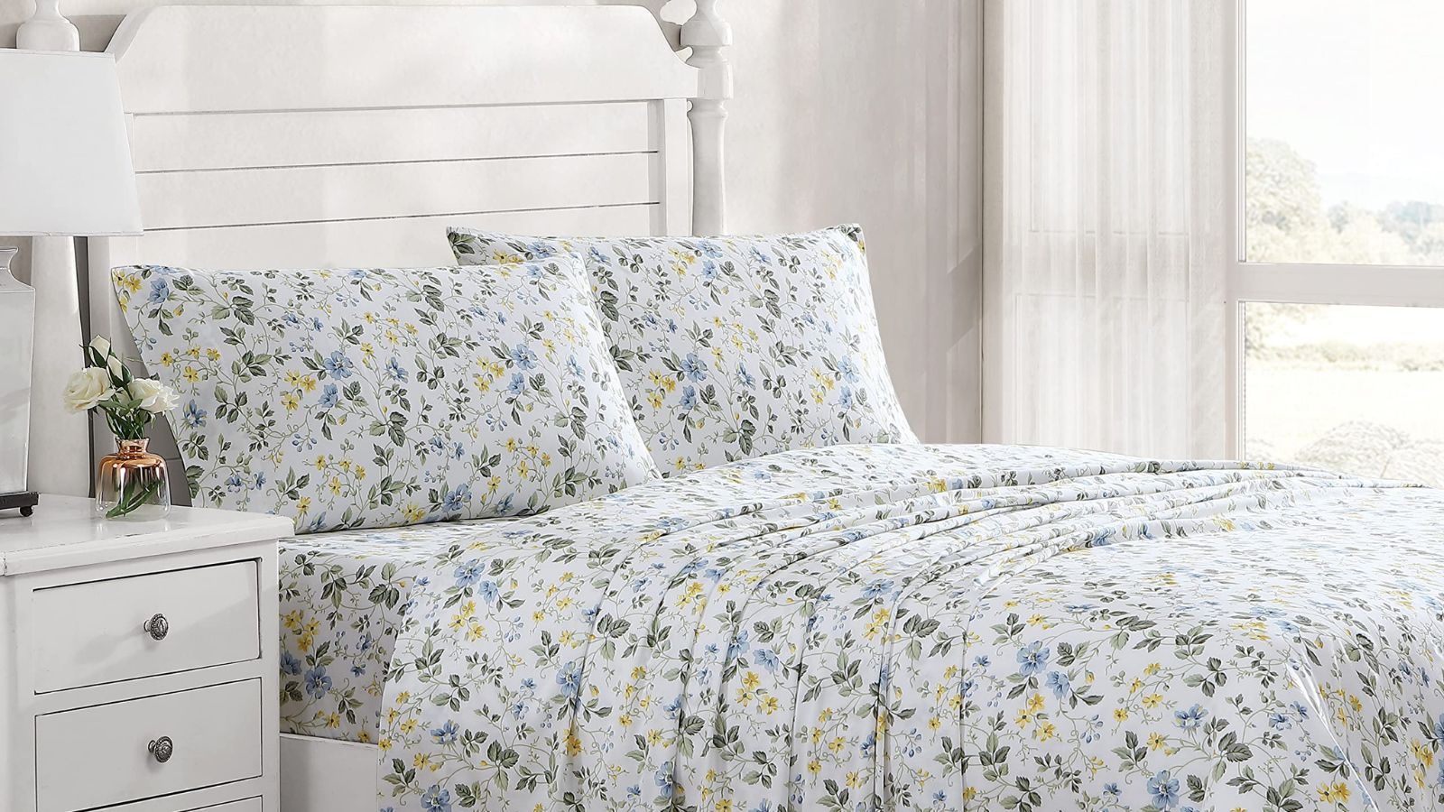 White, green, and yellow floral sheets on a white bed frame against white walls; to the left, a white lamp on a white nightstand; to the right, a sheer curtain covering a window. 