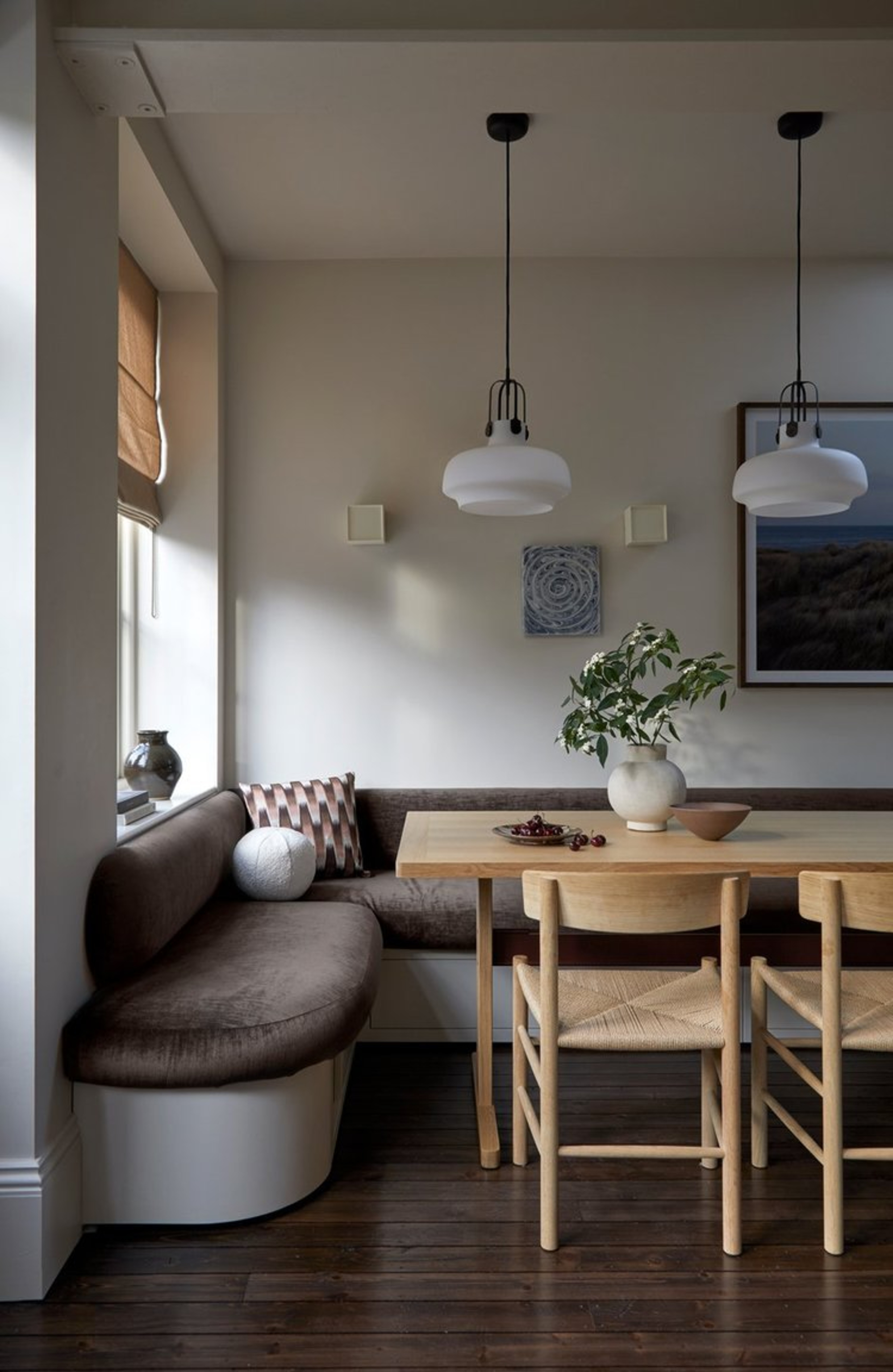 corner of open-plan space with upholstered corner banquette seat, blind over window, long rectangle timber dining table with two seats, two pendant lights hanging above, and a vase with flowers on the table