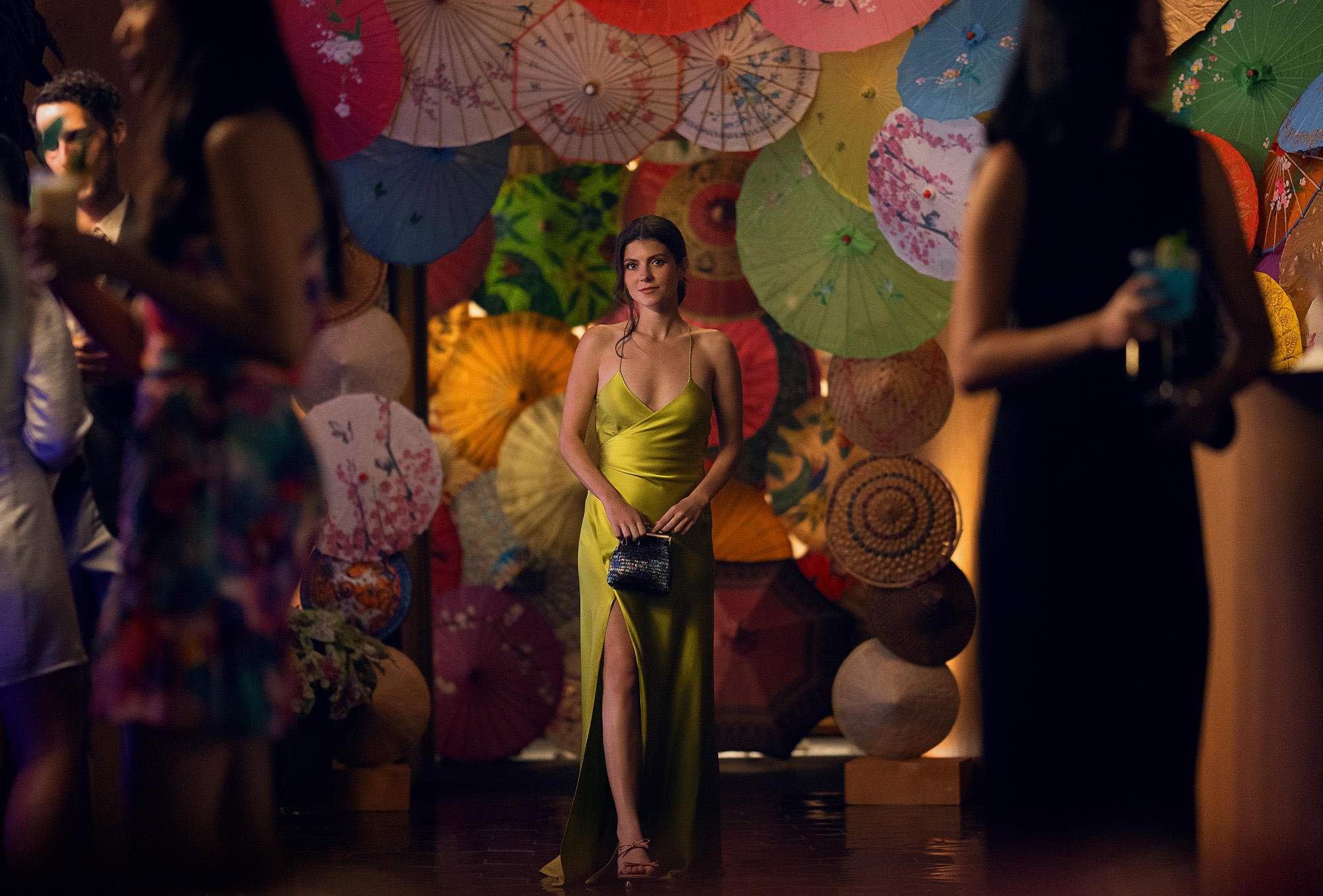 A film still from People We Meet on Vacation featuring character Poppy played by Emily Bader. In the image she is wearing a chartreuse dress with a black clutch.