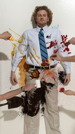 20 year old man lying on the ground wearing a button down shirt, tie and khaki with people pouring ketchup, mustard, salsa, coffee, dirt and wine on a white background