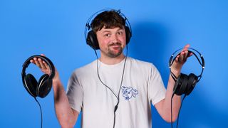 A man holding three pairs of headphones and shrugging at the camera