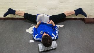a male ballet dancer wearing black athletic pants, a blue adidas zip up jacket, and insulated boots lies on his back reading with his legs propped up in a straddle split against a wall. His head is resting on a foam roller as he reads