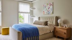 An off-white bedroom with a wood side table, a tiered paper pendant light, a mustard yellow pouf, a blue fuzzy throw, and a beige long pillow