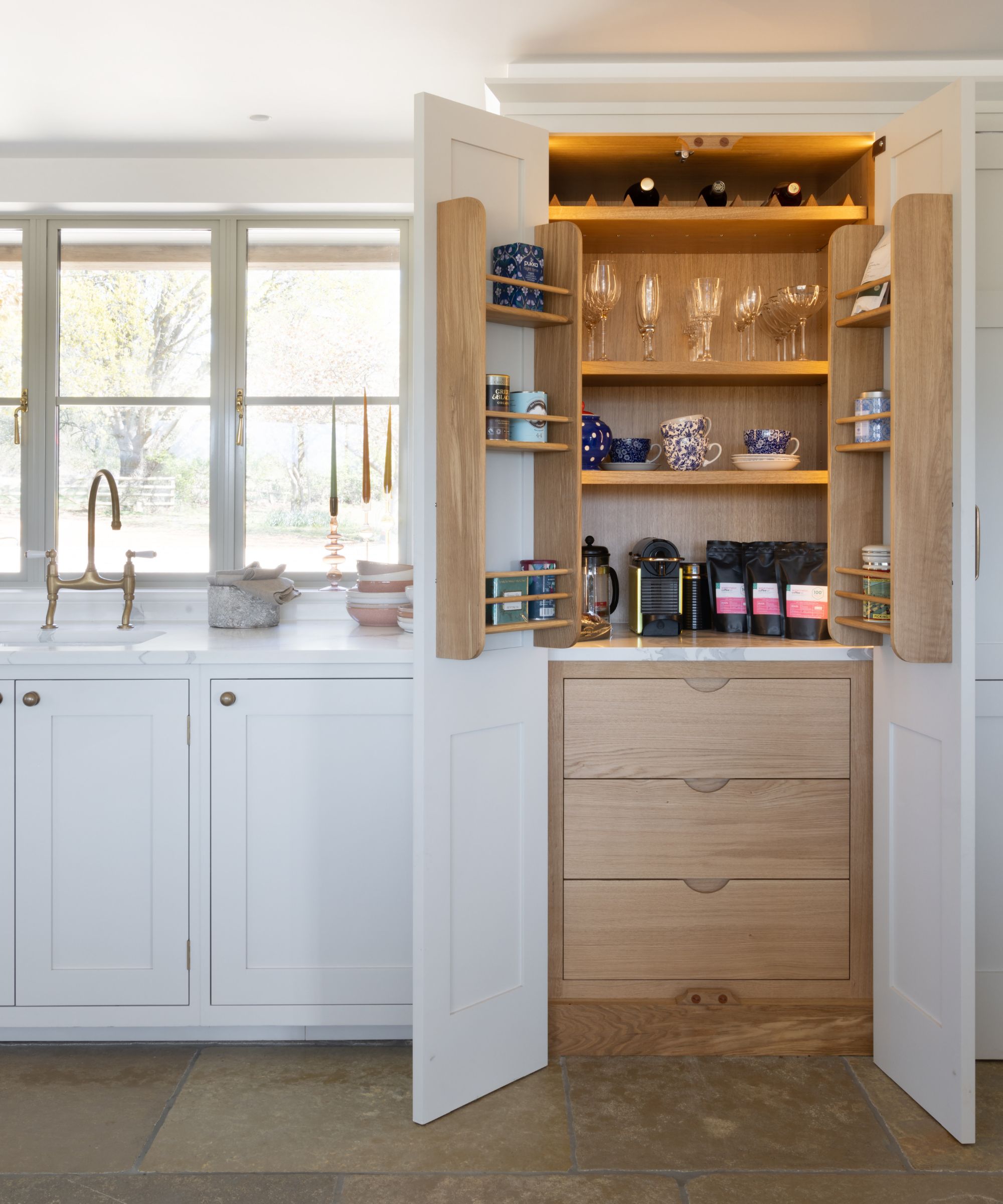 Concealed kitchen coffee station inside a tall cupboard, with open doors revealing shelves for mugs, glasses and coffee equipment.