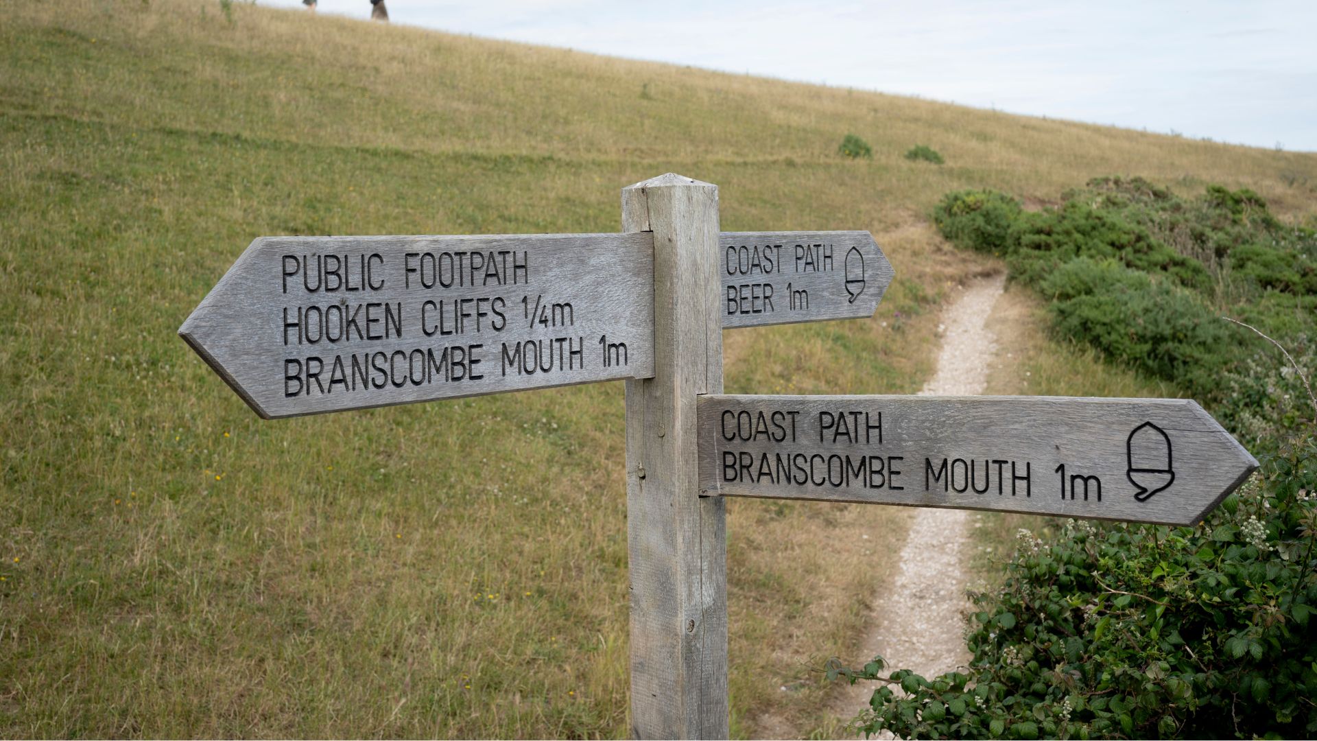 Walkers on a ridge and a signpost gives distances between Branscombe Mouth and Beer on the long-distance coastal path in east Devon, Branscombe