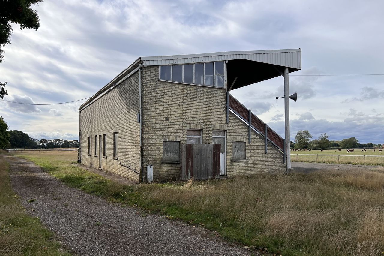 The rear of a horse racing stand shows a brick structure surrounded by overgrown grass