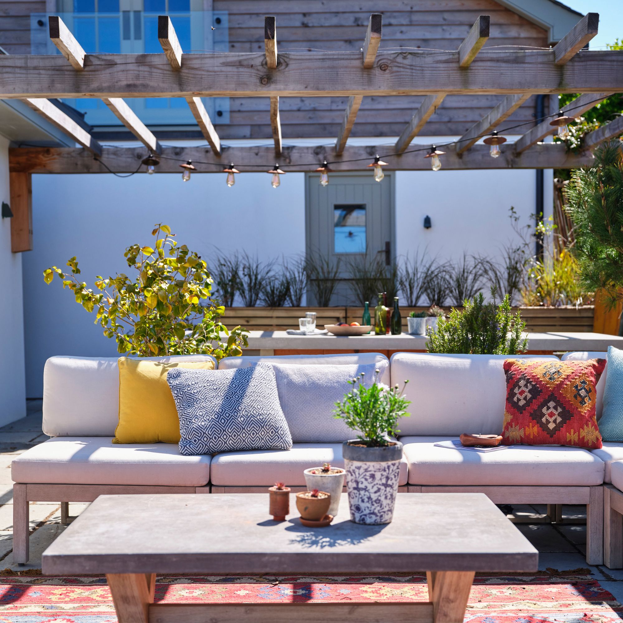 A light wood pergola with string lights above a white furnished garden bench and coffee table