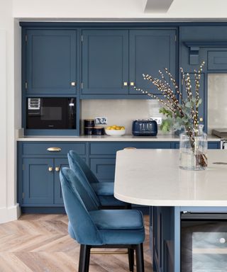 A kitchen with dark blue cabinets, white countertops with a toaster and fruit bowl on it, or a curved white kitchen island with a glass vase with brown stems and two dark blue velvet bar stools