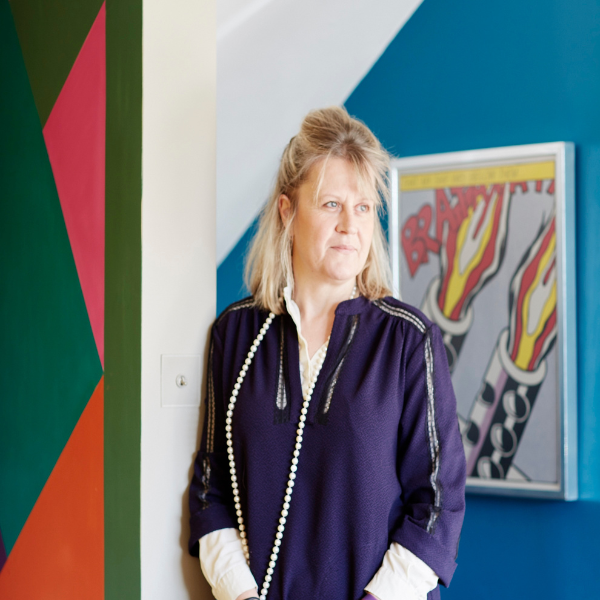 A headshot of Emma Deterding, a lady with shoulder-length blonde hair, half tied up, wearing a navy top with a long white bead necklace, standing against a colorful patterned wall with a blue wall in the background, with a piece of framed pop art hung on it