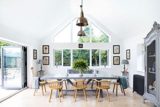 dining space with full height gable end window and open bifold doors pale stone floors gray dining table wooden chairs gray glazed cabinet